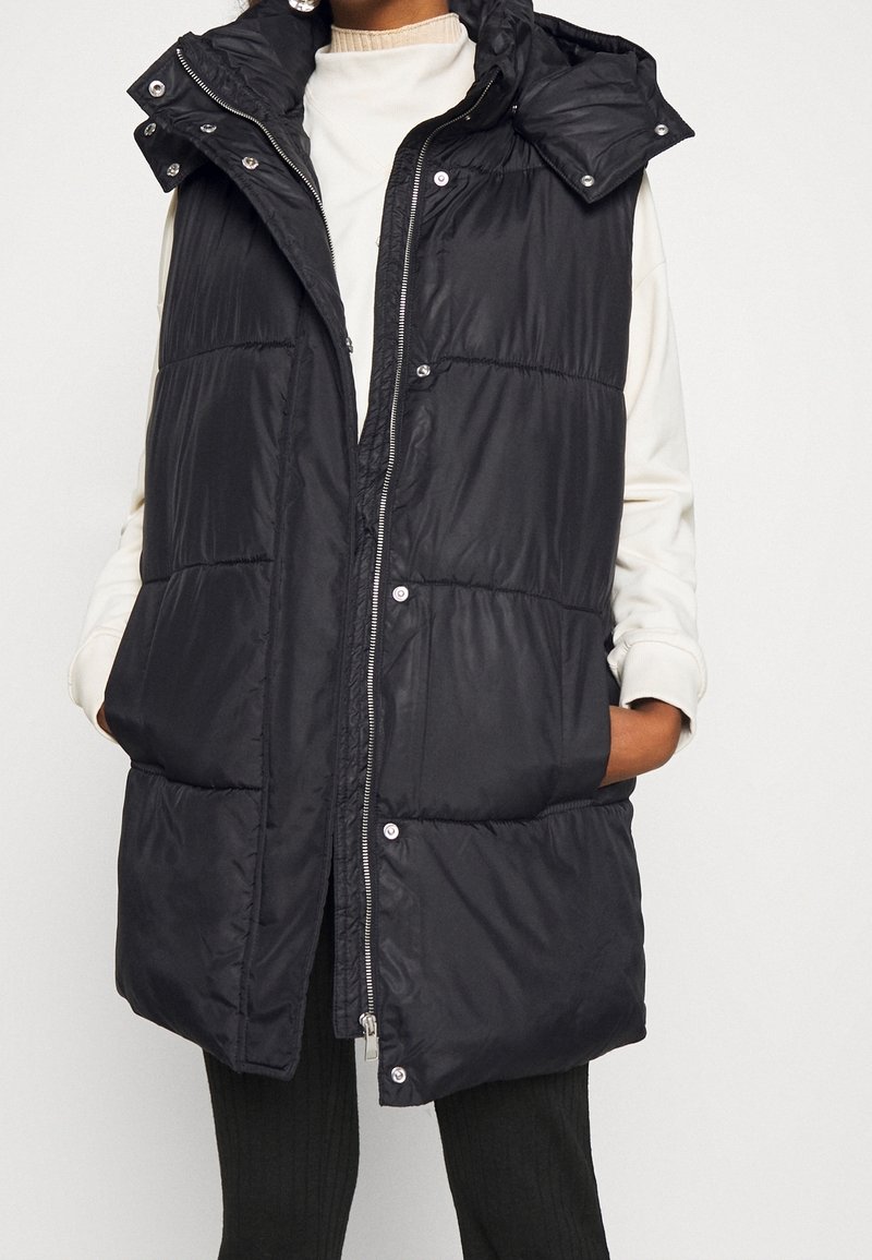 Femme portant un gilet matelassé noir sans manches par-dessus un haut blanc à manches longues, les mains dans les poches du gilet, sur un fond uni.