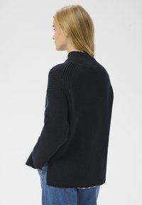 Femme aux longs cheveux blonds portant un pull côtelé foncé et un jean bleu, de profil vers la gauche, sur un fond clair et uni.