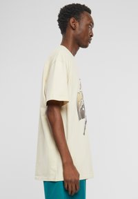 Beige bomulds t-shirt med korte ærmer, der har et grafisk design på fronten. Løs pasform og standard crew neck stil.