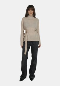 Maglione beige con collo alto e cintura legata, maniche lunghe e vestibilità comoda, abbinato a jeans scuri a gamba dritta. Texture liscia, dettagli minimali.