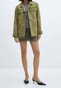 Žena v zelené pracovní bundě přes šedý top, s zelenou krajkovou mini sukní a černými slingback podpatky, stojící na pozadí bez vzoru.