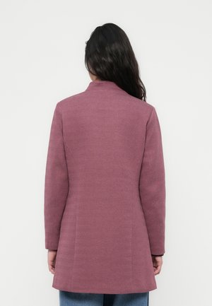 Woman with long dark hair wearing a fitted, textured purple coat standing facing away against a plain light background.