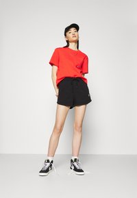 Camiseta roja de manga corta con bolsillo, combinada con pantalones cortos negros con cordón. Zapatillas negras con suelas blancas, usadas con calcetines blancos.