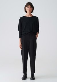 Schwarzes, übergroßes Langarmshirt, kombiniert mit schwarzen, hochgeschnittenen Hosen, die einen lockeren Schnitt und eine glatte Textur aufweisen. Schwarze Knöchelstiefel.