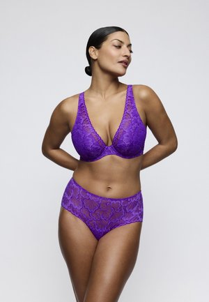 Woman with dark hair in a bun wearing matching purple floral lace bra and high-waist underwear, standing with hands behind her back.