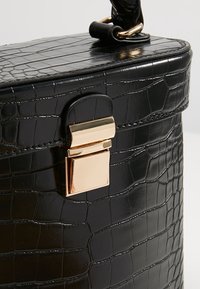 Coin de sac à main noir à texture crocodile avec fermoir rectangulaire doré et détail de poignée tressée sur un fond clair.