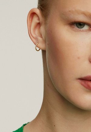 Aro de oro con una fila de pequeñas piedras preciosas transparentes, situado sobre un tono de piel claro y un fondo suave y neutro.