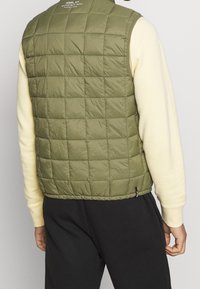 Olivgrön vadderad väst med hög krage, tillverkad av återvunnen nylon, med en diskret logotyp på baksidan och en rak nederkant.