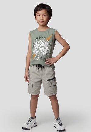 Niño de pie con la mano en la cadera, llevando una camiseta sin mangas verde con estampado de tigre, pantalones cortos cargo beige y zapatillas blancas sobre un fondo gris.