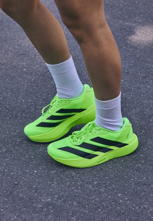 Baskets de course vert lime avec des rayures noires, tige en maille et semelle épaisse. Portées avec des chaussettes blanches, reposant sur une surface en asphalte gris.