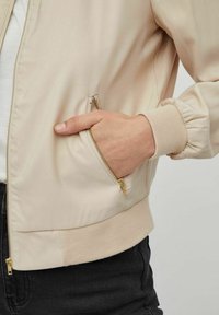 Beige bomber bunda s ribovanými manžetami, bočním zipovým kapsou, hladkým texturovaným fabricem a viditelnými zlatými kovovými doplňky. Klasický zkrácený design.