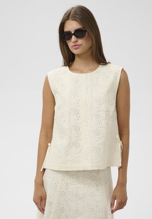 Junge Frau trägt schwarze Sonnenbrille und ein ärmelloses cremefarbenes Eyelet-Top mit passendem Rock vor einfarbigem Hintergrund.