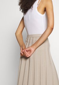 Beige Faltenrock aus weichem Stoff; kombiniert mit einem enganliegenden weißen Tanktop. Der Rock verfügt über vertikale Streifen und einen hohen Bund.