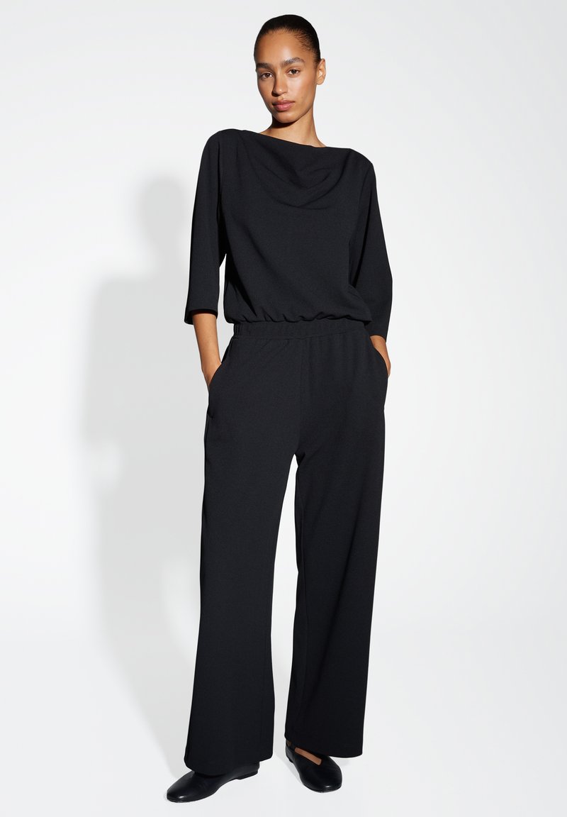 Tuta nera con scollatura drappeggiata, maniche 3/4, vita elasticizzata e design a gamba larga. Texture liscia e stile minimalista.