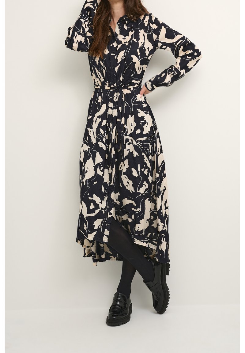 Langes, marineblaues Kleid mit einem cremefarbenen abstrakten Muster, durchgehender Knopfleiste, langen Ärmeln und einem Gürtel in der Taille, kombiniert mit schwarzen Schuhen und Strumpfhosen.