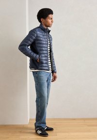 Blå puffjakke med horisontale sømmer, kombinert med en svart-hvit stripete skjorte, lyseblå jeans og svarte sneakers med hvite detaljer.