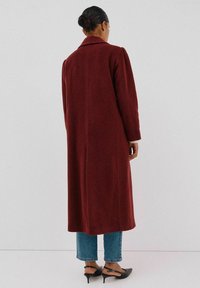 Lange burgundy jas in een wolmix, met een geknipte kraag, gestructureerde schouders en een rechte snit, gecombineerd met blauwe jeans en hakken.