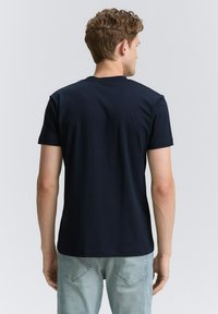 Tricou bleumarin cu manșetă scurtă, dintr-un material de bumbac neted; guler rotund și o croială lejeră. Vedere din spate, asortat cu blugi de un albastru deschis.
