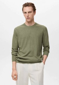 Olive green knit sweater with long sleeves, round neckline, textured weave, and relaxed fit; paired with light-colored trousers.