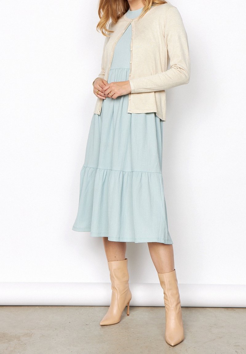Femme portant une robe midi bleu clair à étages, un cardigan beige boutonné et des bottines beiges pointues à talons, debout sur un sol gris.