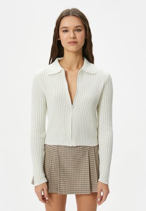 Cardigan a coste in avorio con ampio colletto, chiusura con zip e maniche lunghe, abbinato a una mini gonna a quadri in toni di marrone.