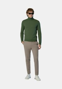 Maglione verde a collo alto in materiale lavorato a maglia, abbinato a pantaloni beige e sneakers bianche. Il modello indossa occhiali da sole neri.