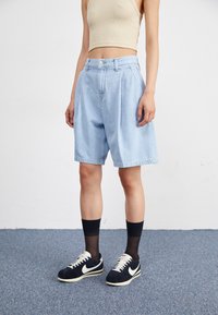 Shorts en denim bleu clair avec une coupe décontractée, dotés d'une taille haute et d'un devant plissé. Assortis avec des chaussettes noires et des baskets sombres.