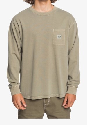 Mężczyzna w oliwkowej koszuli z długim rękawem z małą kieszonką na piersi i pasujących spodniach, stojący na białym tle.