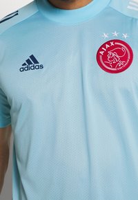 Jersey deportivo Adidas de color azul claro con un diseño de malla texturizada, que presenta un logo rojo de Ajax y tres estrellas blancas encima.