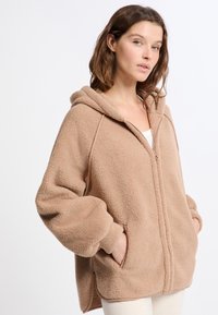 Felpa in pile color sabbia con zip frontale, grandi tasche e una vestibilità ampia e comoda. Presenta un cappuccio con coulisse e dettagli a costine sui polsini.