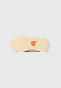 Suela de zapato en goma de color crema claro con un patrón de banda de rodadura en zigzag; un logo naranja está centrado en la base. Textura suave y forma redondeada.