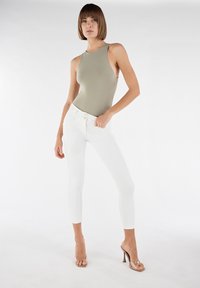 Bodysuit sans manches vert olive associé à un pantalon skinny blanc ; sandales à talons blocs transparentes, textures lisses et coupe ajustée.