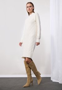 Robe pull en cream avec tricot torsadé, manches longues, poignets côtelés et coupe droite ; associée à des bottes en daim beige à talons aiguilles, montantes jusqu'au genou.
