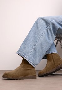 Persona vestida con jeans azul claro y botines de ante marrón, sentada en una silla con las piernas cruzadas.
