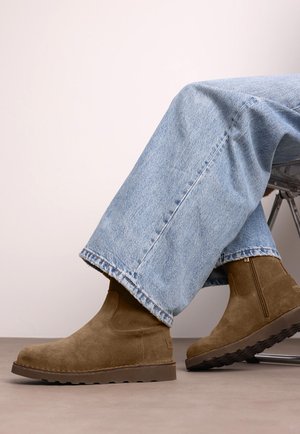 Persona che indossa jeans azzurri chiari e stivaletti in suede marrone mentre è seduto su una sedia con le gambe incrociate.