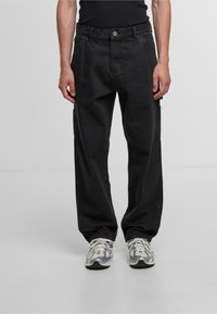 Zwarte denim cargobroek met een relaxte pasvorm, voorzien van zijzakken, een ritsdetail en contrasterende stiksels. Gepaard met grijze sneakers.