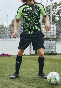 Svart och neongrön atletisk tröja med ett bladmönster, ihop med svarta shorts och knähöga strumpor, på en fotbollsplan.