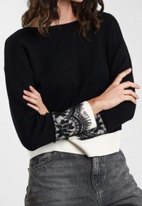 Maglione nero con orlo inferiore bianco sporco e dettagli in pizzo sui polsini. Tessuto morbido, vestibilità rilassata e maniche lunghe.