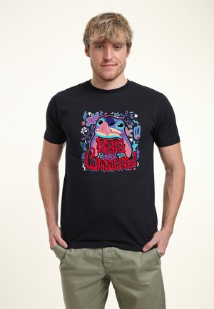 Jeune homme aux cheveux blonds portant un t-shirt noir avec un chien de dessin animé coloré et le texte « Weird and Complicated », assorti à un pantalon vert clair.
