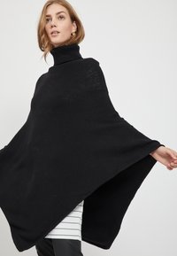 Poncho preto com gola alta. Tecido de malha suave; design largo e fluido com aberturas para os braços. Sobreposto a uma peça listrada.