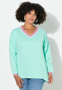 Donna con capelli ricci che indossa un top a maniche lunghe verde menta con scollatura a V a righe rosse e bianche e polsini, abbinato a jeans blu, in piedi contro un muro bianco.