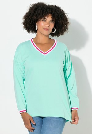 Kvinde med krøllet hår iført en mintgrøn langærmet top med rød- og hvidstribet V-hals og manchetter, parret med blå jeans, stående mod en hvid væg.