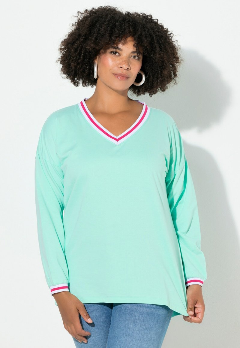 Donna con capelli ricci che indossa un top a maniche lunghe verde menta con scollatura a V a righe rosse e bianche e polsini, abbinato a jeans blu, in piedi contro un muro bianco.