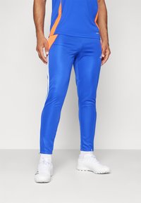 Pantalon de sport bleu avec une coupe ajustée, présentant des accents latéraux orange et des rayures blanches sur les côtés. Fabriqué dans un matériau lisse et extensible.