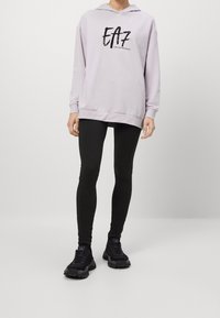 Felpa viola chiaro con cappuccio e logo EA7 nero, abbinata a leggings neri e sneakers nere chunky. Presenta una vestibilità rilassata e maniche lunghe.