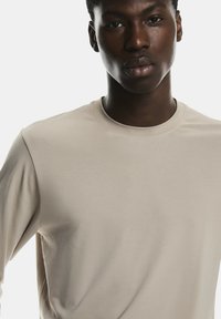 Cămașă bej cu mânecă lungă dintr-un material moale, cu decolteu rotund. Textură netedă, design minimalist, siluetă ajustată și fără modele sau accente vizibile.