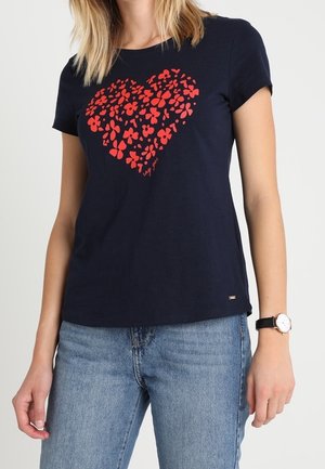 Vrouw draagt een marineblauw T-shirt met rood bloemenhartontwerp en lichtblauwe jeans, aangevuld met een zwarte polshorloge.