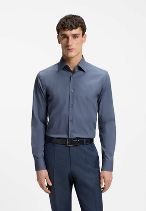 Man wearing a buttoned-up dark blue dress shirt tucked into navy trousers with a black belt, standing against a plain white background.