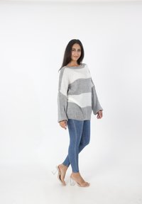 Grijs en wit gestreepte oversized trui met brede mouwen, gecombineerd met slanke blauwe jeans en doorzichtige hakken. Witte achtergrond.