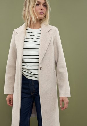 Manteau beige clair avec des revers structurés et un bouton unique. Porté par-dessus un top rayé blanc et noir, associé à un jean foncé.